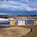 Biało-niebieski napis Darlowo przy deptaku na tle morza